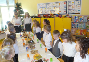 Pani dyrektor Maria Królikowska, pani Ewa Janicka oraz dzieci stoją wokół nakrytego białym obrusem i poczęstunkiem stołu.